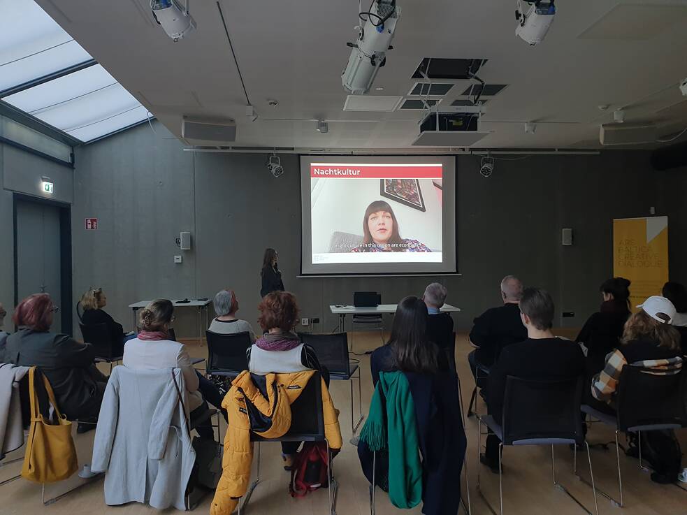 Presentation scene in a room with attendees seated and facing a speaker standing beside a screen. The slide on display features an image. The room includes overhead lighting and large windows