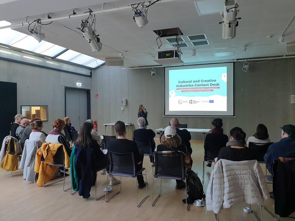 Photo of a presentation in progress, showing attendees seated. A speaker stands near a screen displaying the title 'Cultural and Creative Industries Contact Desk' alongside logos of the European Union and other organizations.