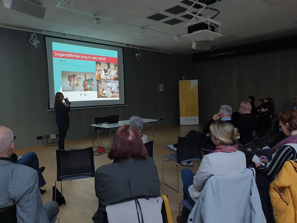 Presentation scene with attendees seated and facing a speaker standing near a screen. The slide on display is titled Jugendförderung in der KKW (Youth Promotion in the KKW) and features images of children participating in various activities