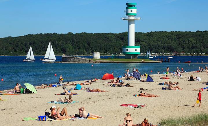 Ein Sandstrand am See, Personen liegen im Sand und sonnen sich. Im Wasser sieht man Segelboote, ein Leuchtturm ist in der Mitte des Sees.