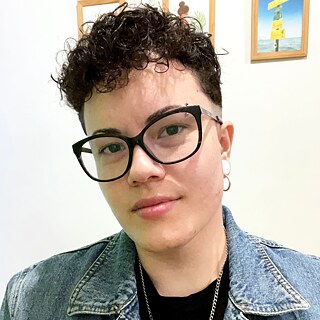 Suellen Scanavini, mediadora da Oficina. Pessoa com cabelo cacheado, usando óculos e brincos, vestindo jaqueta jeans sobre camiseta preta. Ao fundo, duas imagens emolduradas na parede.