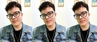 Retrato da Suellen Scanavini, mediadora da oficina de stop-motion. Mulher com cabelo cacheado curto, usando óculos e brincos, vestindo jaqueta jeans sobre camiseta preta. Ao fundo, duas imagens emolduradas na parede.