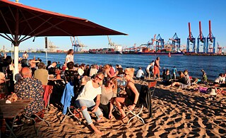 Personen sitzen auf Stühlen an einem Sandstrand am Fluss "Elbe". Auf der anderen Seite des Flusses sieht man Containerschiffe und große Hafenkräne.
