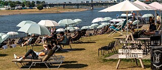 Sonnenliegen und Sonnenschirme am Ufer eines Flusses. Ein Schild mit der Aufschrift "Willkommen am Stadtstrand"