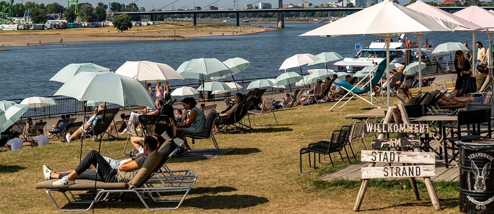 Sonnenliegen und Sonnenschirme am Ufer eines Flusses. Ein Schild mit der Aufschrift "Willkommen am Stadtstrand"