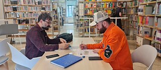 Zwei Männer sitzen an einem Tisch in einer Bibliothek und arbeiten gemeinsam an einem Arbeitsblatt. Im Hintergrund sind Bücherregale und eine weitere Person zu sehen.