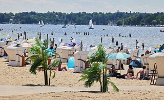 Ein Sandstrand mit Strandkörben an einem See. Im Wasser fahren Segelboote, am Strand sitzen Personen in Badekleidung.