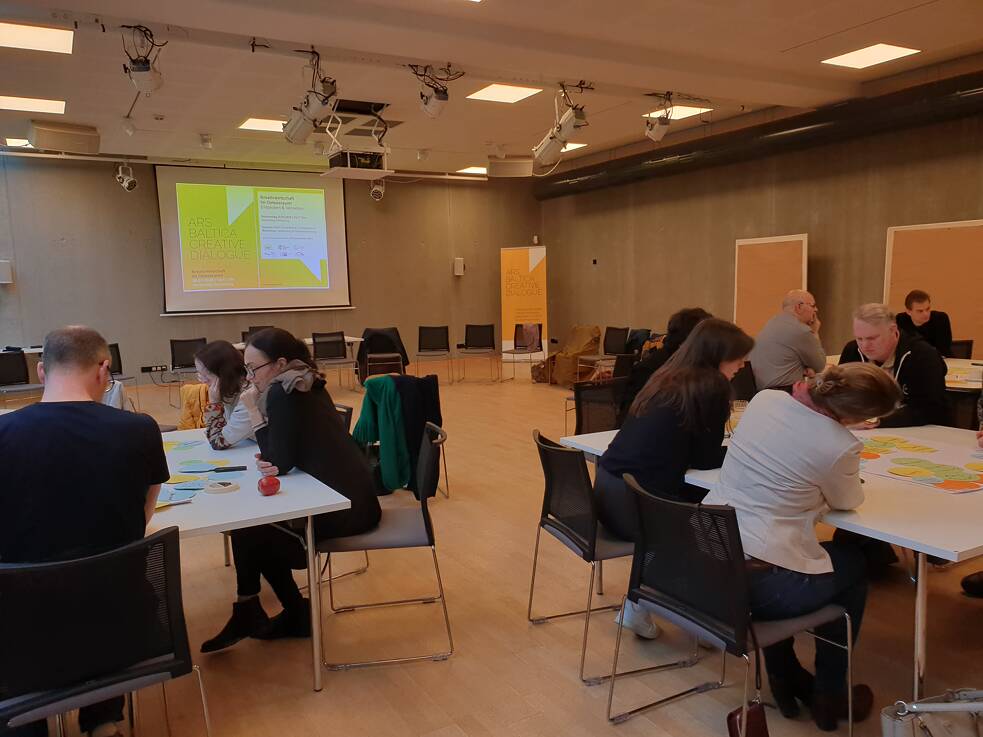 Conference or meeting room with participants seated at tables, facing a large screen displaying a presentation slide with text and graphics. The room includes posters or boards on the walls and chairs arranged in rows. Attendees appear to be engaged in discussions or activities related to the presentation.