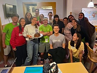 Eine Gruppe feiert Geburtstag; eine junge Frau hält einen Gugelhupf mit einer Kerze in der Hand.