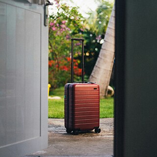 Ein roter Hartschalenkoffer mit Rollen steht auf einer Betonfläche direkt vor einer offenen Tür, umrahmt von einem üppigen grünen Garten mit Palmen und leuchtenden Blumen im Hintergrund.