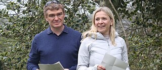 Zwei Alumni des Europanetzwerk Deutsch stehen mit Papieren in der Hand nebeneinander vor einem grünen Busch.