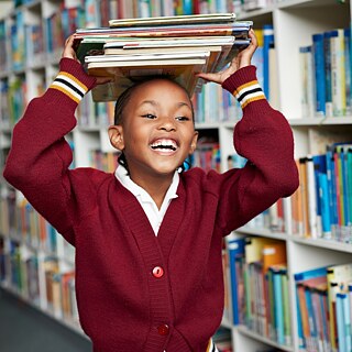 Eine Schülerin in der Bibliothek. Das Mädchen steht zwischen den Regalen und balanciert einen Stapel Bücher auf dem Kopf, sie lacht.