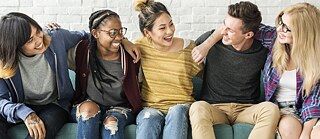 Teenagers are sitting on a sofa in front of a white stone wall and laughing together.