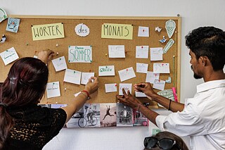 A Summer Academy participant and a language course participant hang cards on a bulletin board for the words of the month