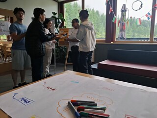 Ein buntes, selbst gemaltes Plakat liegt auf einem Tisch; im Hintergrund eine kleine Gruppe Jugendlicher.