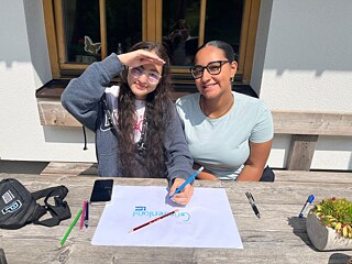 Jugendliche sitzen im Freien an einem Tisch und malen ein Plakat.