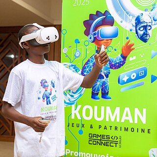 Ein Foto eines Jugendlichen mit VR-Brille vor einem Kouman-Banner bei einem Kouman-Workshop in Yamoussoukro.