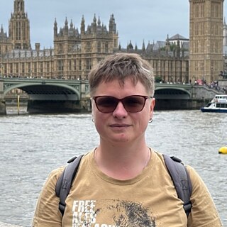 Eine Frau mit beigem T-Shirt und Sonnenbrille steht vor der Themse