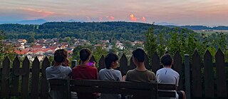 Jugendliche auf einer Parkbank blicken bei Sonnenuntergang auf eine Stadt, oder auf ihr Smartphone.