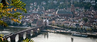 View of Heidelberg’s Old Bridge over the Neckar River, framed by green and yellow leaves. © © Unsplash Bridging Cultures, Building Futures