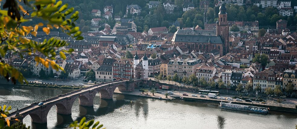 View of Heidelberg’s Old Bridge over the Neckar River, framed by green and yellow leaves.