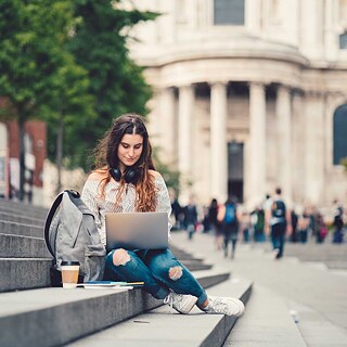 Eine junge Frau sitzt auf den Treppenstufen, auf dem Universitätsgelände und lernt am Laptop.