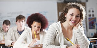  Mehrere Schüler sitzen bei einer Prüfung im Klassenzimmer. Die Schülerin im Vordergrund lächelt in die Kamera.