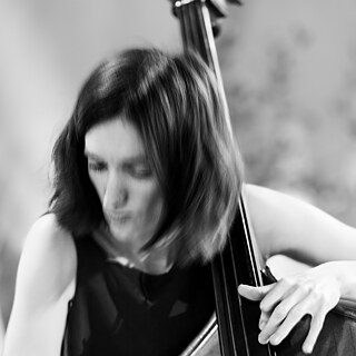 Das Bild ist ein künstlerisches Schwarz-Weiß Porträt der Musikerin Natasha Zaychenko, auf dem nur ihre Hand auf dem Kontrabass im Fokus ist, während ihr Kopf und Gesicht im Hintergrund verschwommen zu erkennen sind.