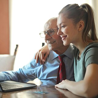 Eine junge Frau zeigt einem älteren Mann etwas auf einem Laptop.
