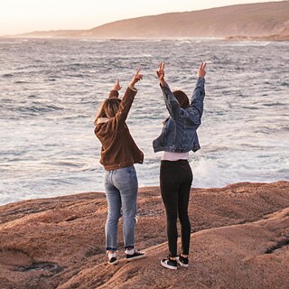 Zwei Mädchen strecken am Meer die Hände in die Luft.