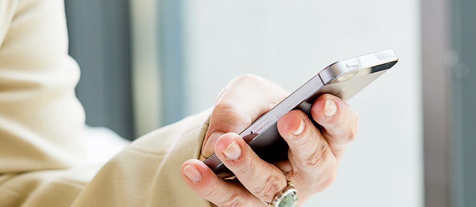 Eine Person in einem beigen Blazer hält ein Smartphone in der einen und eine Zeitung in der anderen Hand. Auf dem Tisch liegt eine weitere Zeitung. Im Hintergrund große Fenster.