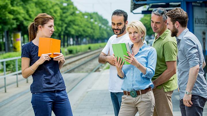 A group of five people is standing on a tram or train platform. One person is holding an orange book, while two others are looking at a green tablet. In the background, there are tracks, trees, and a blue sign that reads “Goethe-Institut.”