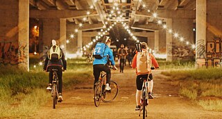 Three young man on their bikes from behind passing under a lit up bridge