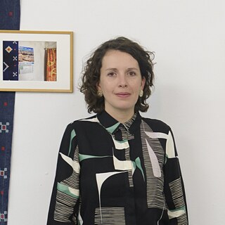 Mariana Sesma steht einer Ausstellungseröffnung neben einem Kunstwerk vor einer weißen Wand.