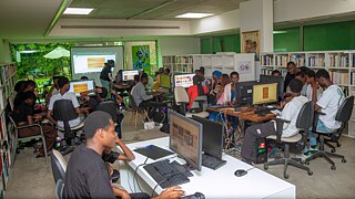 Kollaborativer digitaler Workshop bei Kouman 2025 in Abidjan, bei dem die Teilnehmer in einem hellen, mit Büchern ausgestatteten Bibliotheksraum mit einer großen Präsentationsleinwand an Computern arbeiten.