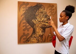 Kunstbetrachtung bei Kouman 2025 in Abidjan, wo ein Teilnehmer ein detailliertes Seitenprofilgemälde in Gold- und Brauntönen fotografiert und damit den kreativen und kulturellen Schwerpunkt der Veranstaltung hervorhebt.