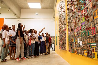 Studierende interagieren mit einer lebendigen Kunstinstallation zum Thema Architektur bei Kouman 2025 in Abidjan, vor einer leuchtend gelben Galeriewand in einem modernen Ausstellungsraum.