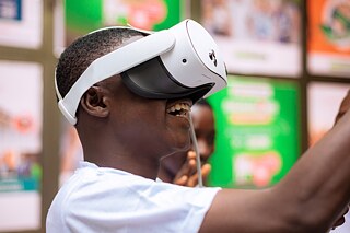 A smiling Kouman workshop participant wearing VR glasses with his arms in the air. 