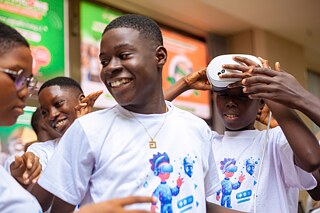 A group of Kouman participants wearing matching white t-shirts with a colorful graphic of a person using virtual reality technology, engaging in an interactive experience with a VR headset.