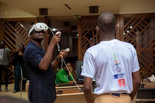 A Kouman participant is using a virtual reality headset and controllers in an indoor space with other participants engaged in technology-related activities, surrounded by computer equipment.
