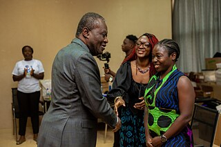 A group of Kouman participants gathered indoors, engaging in conversation and handshakes. The individuals wear a mix of traditional and formal attire.