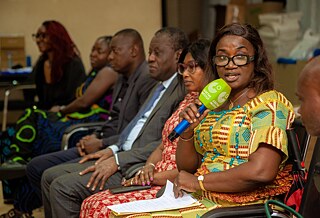 A group of seated Kouman participants, with one individual speaking into a microphone labeled "Goethe Institute." The attendees are dressed in formal and traditional attire. 