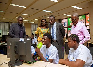 A group of Kouman workshop participants, with some seated at desks using computers and others standing and observing. One person holds a microphone labeled "Côte d'Ivoire," suggesting an presentation. 
