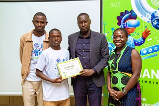 Group photo from a Kouman event in 2025, showcasing four participants standing together. One individual holds a certificate of participation, with a vibrant digital-themed banner in the background highlighting the year 2025.