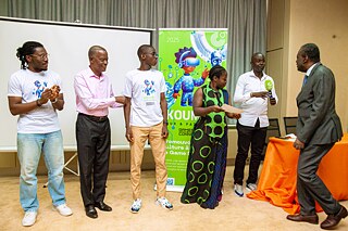 Award presentation at Kouman 2025, with participants in front of a colorful banner promoting culture through games.