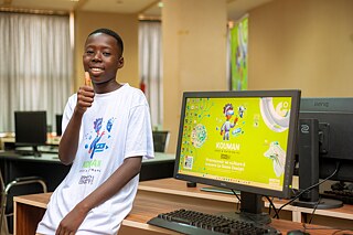 A Kouman workshop participant giving a thumbs up, standing in front of a monitor showing Kouman workshop related content. 