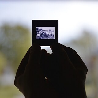 Zwei Hände im Gegenlicht halten ein Dia, das eine Gruppe von Dorfbewohner*innen zeigt