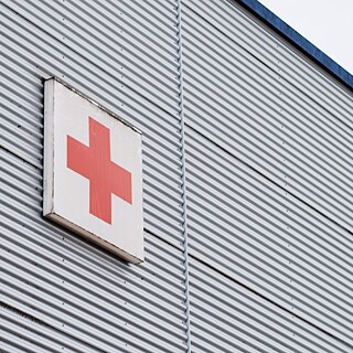 Man sieht den oberen Teil eines grauen Gebäudes mit einem Schild, das ein rotes Kreuz auf weißem Grund zeigt.