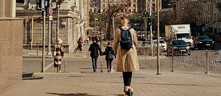 Stadtstraße mit Fußgängern und einer Person auf einem E-Scooter mit beigem Mantel und blauem Rucksack.