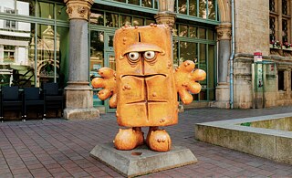 A statue of a loaf of bread with hands, feet and a grim face stands in front of a building with many glass windows.